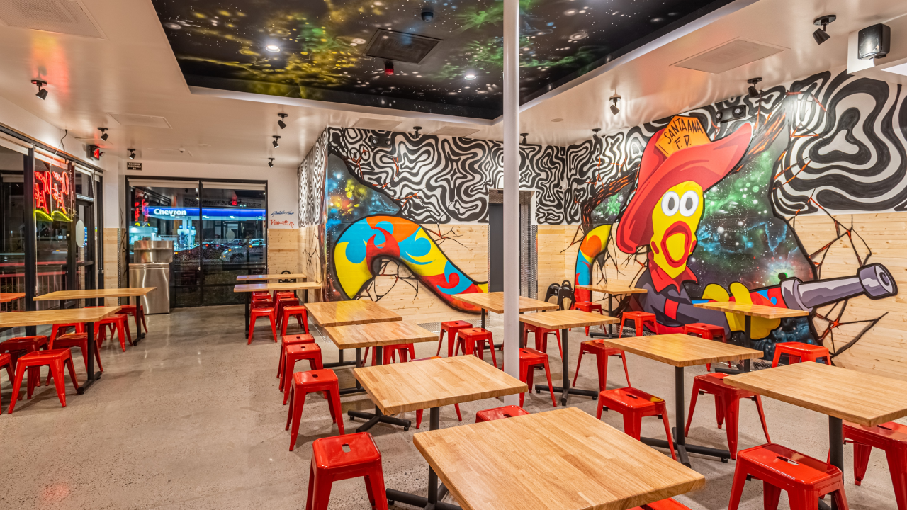 Interior image of Dave's Hot Chicken in Santa, Ana. Focusing on the dining area with wooden tables and red chairs. In the background you can see the fried chicken restaurant in Santa Ana's signature graffiti.