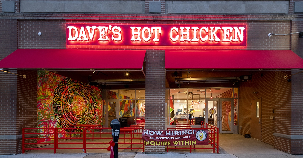 Exterior photograph of a Dave's Hot Chicken Restaurant on Detroit Avenue at night. The bright red neon sign and colorful hand painted mural inside is almost as inviting as thier Nashville style hot chicken.