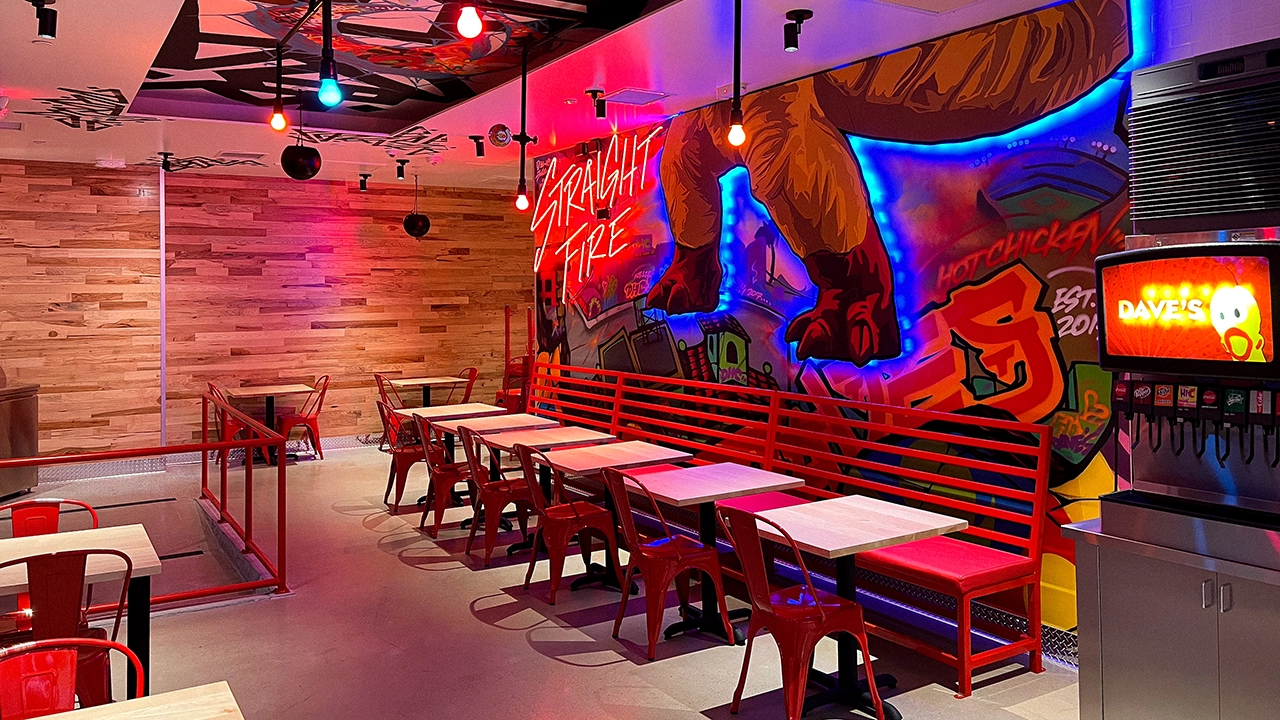 The dining area of Dave's Hot Chicken in Little Tokyo, featuring a colorful wall mural with 'STRAIGHT FIRE' in neon lights, red chairs, and wooden tables.