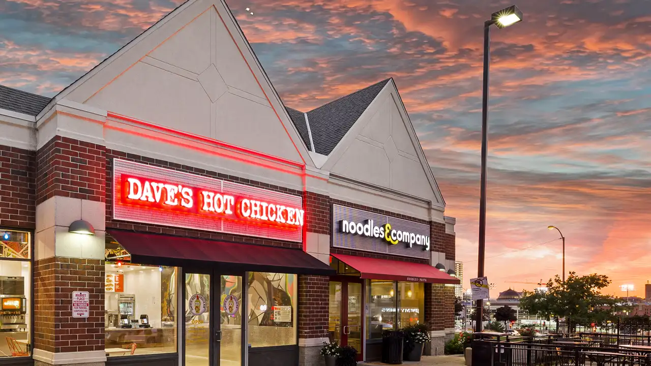 A vibrant evening view of Dave's Hot Chicken restaurant located on Ogden Avenue in Milwaukee, Wisconsin, showing its brightly lit red neon sign. Adjacent is Noodles & Company, under a striking sunset sky with shades of pink and orange. The buildings feature classic brick construction with modern lighting, enhancing the urban setting.