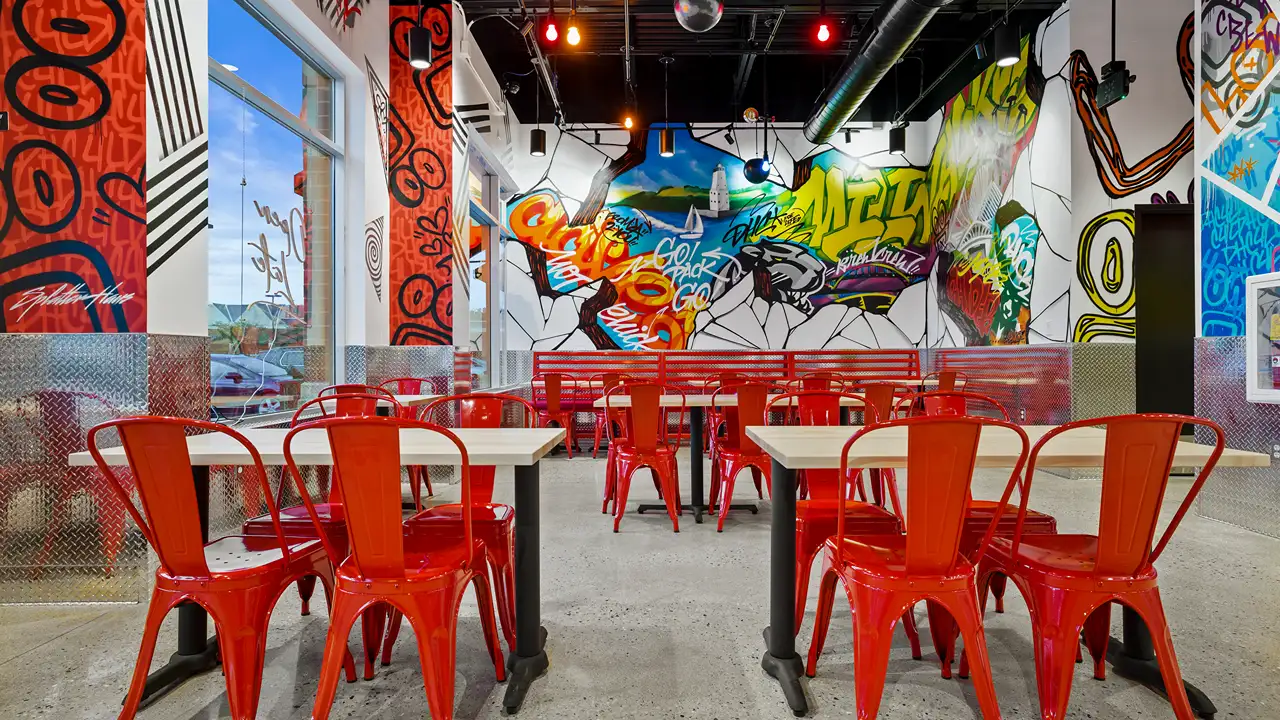 Interior of Dave's Hot Chicken restaurant featuring a vibrant, artsy decor. The room is filled with bright red metal chairs and white tables, set against colorful graffiti-style wall murals depicting urban and abstract themes, including phrases like 'Go Pack Go'. Large windows allow natural light to accentuate the bold colors and energetic atmosphere of the space.