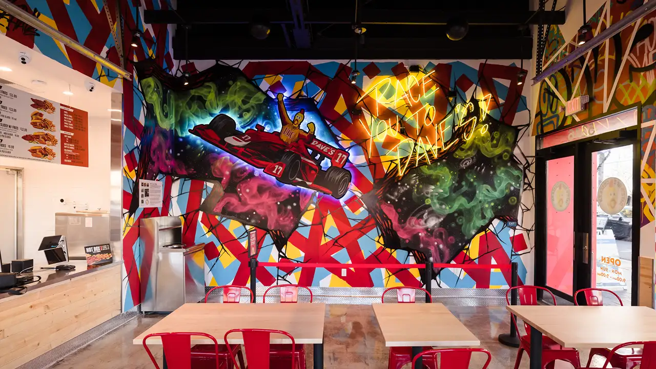Interior of Dave's Hot Chicken restaurant featuring an artistic and colorful mural with a racing theme. The mural shows a red race car labeled 'Dave's 13' driven by a cartoon chicken, surrounded by vibrant cosmic and abstract patterns. The words 'Race you to Dave's!' are illuminated in neon lights above the mural. The dining area includes red metal chairs and wooden tables, with menu boards on the walls.