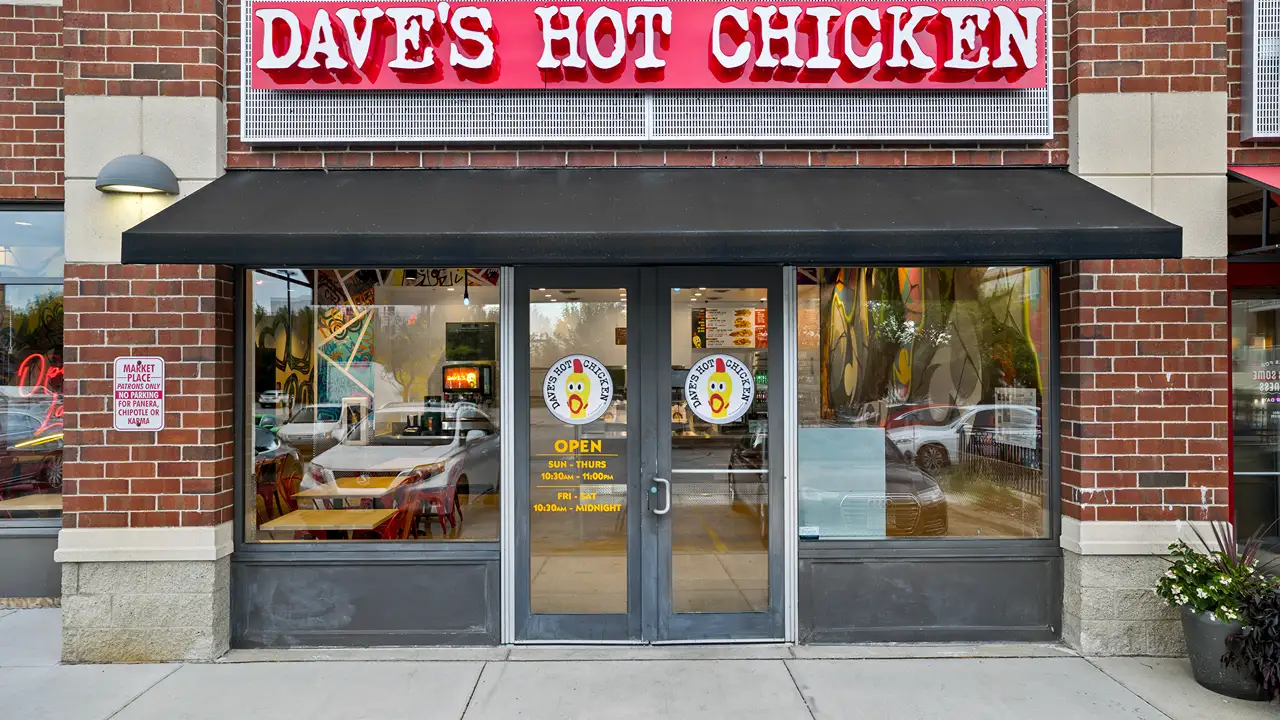 Front entrance of Dave's Hot Chicken restaurant, featuring a bold red neon sign above black framed glass doors. The exterior is constructed of traditional red brick with a small sign on the brick column indicating 'Marketplace No Parking Except for Pickup or Catering.' Reflective windows offer a glimpse of the vibrant interior design and seating area.