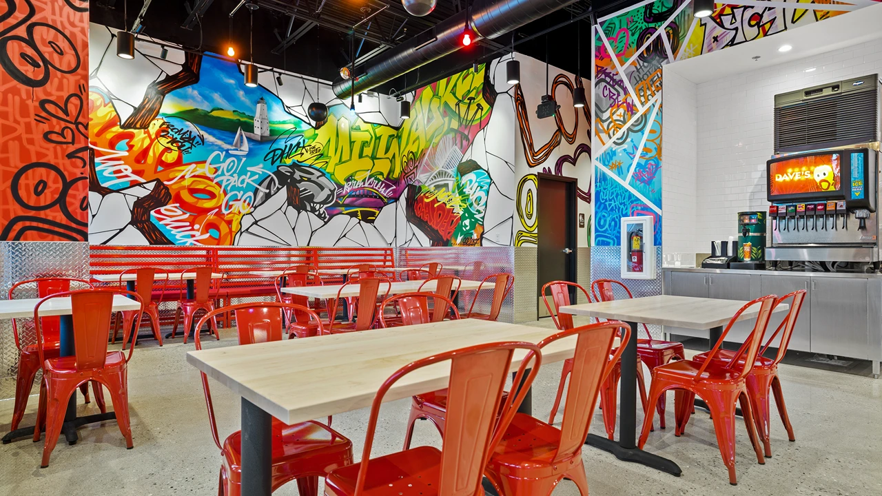 Interior view of Dave's Hot Chicken restaurant showcasing a lively dining area with bright red metal chairs and light wood tables. The walls are adorned with colorful, graffiti-inspired murals featuring dynamic scenes and expressions like 'Go Pack Go'. A serving station with soda dispensers is visible in the background, enhancing the casual, urban vibe of the space.