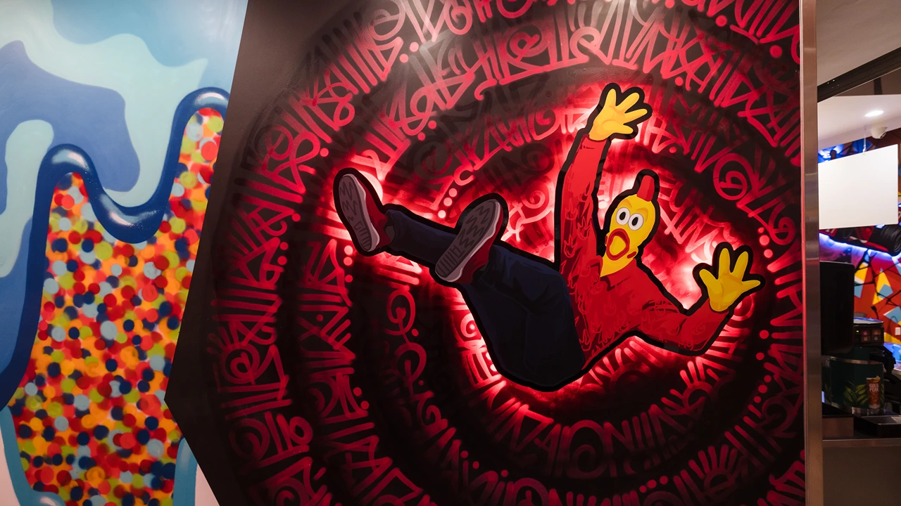 Vivid mural inside Dave's Hot Chicken restaurant depicting a whimsical cartoon chicken caught in a swirl of red with dynamic white script patterns in the background. The chicken, with a surprised expression, is wearing a red outfit and yellow gloves, appearing as though it is tumbling through the swirl.