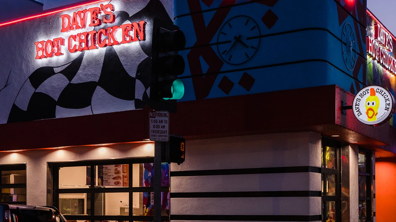 Night view of Dave's Hot Chicken in Long Beach, California. The restaurant's exterior features striking neon red signage spelling out 'Dave's Hot Chicken' against a dark background with abstract black and white patterns. The building is painted in vibrant blue with a large clock and colorful abstract art, and a circular logo of a cartoon chicken is displayed on a corner. A traffic light showing green adds a dynamic urban element to the scene.