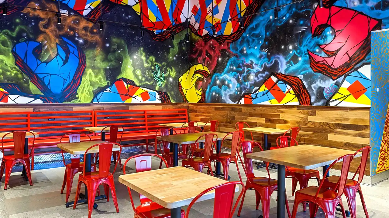 Interior of Dave's Hot Chicken in Palm Desert, California, featuring vibrant and colorful cosmic-themed mural artwork on the wall. The space is furnished with simple wooden tables and bright red metal chairs, creating a lively and casual dining atmosphere.