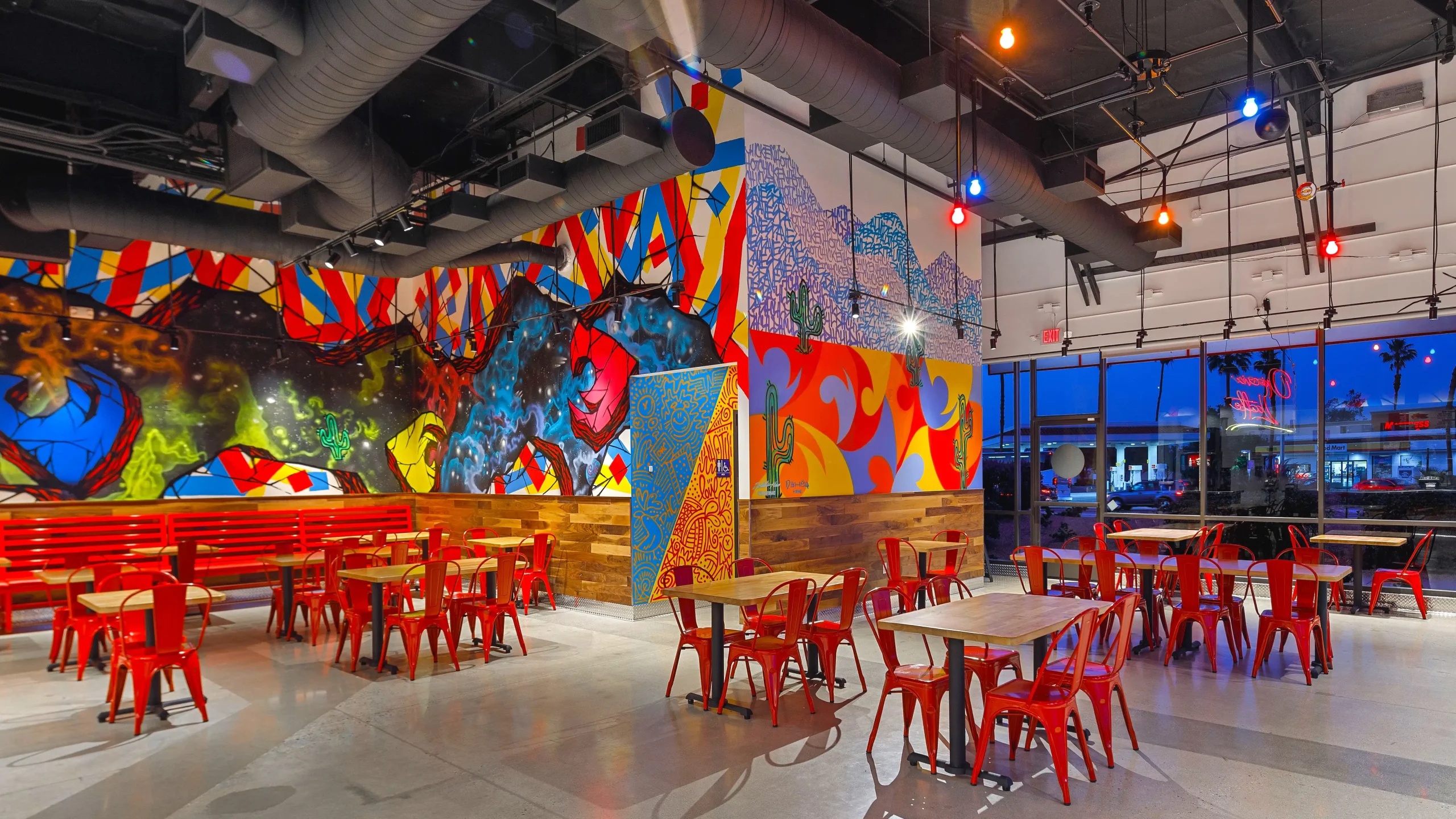Wide view of the interior of Dave's Hot Chicken in Palm Desert, California, showcasing an expansive, colorful mural with abstract and cosmic designs on the main wall. The dining area features sleek wooden tables and vivid red chairs under eclectic, colorful hanging lights. Large windows offer a view of the street outside, illuminated by the evening lights.
