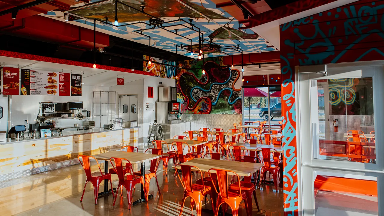 Bright seating area with orange chairs and tables beneath a mural and overhead menu board at Dave's Hot Chicken in Fargo, ND.