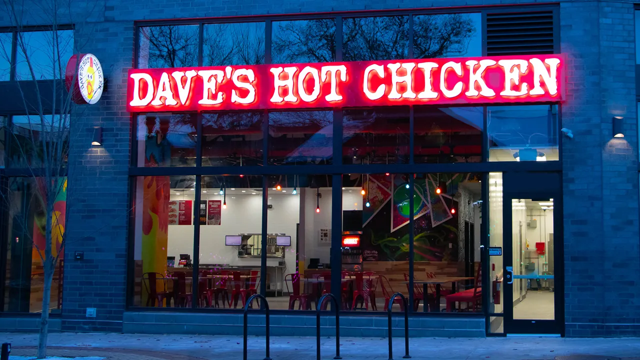 Night view of Dave's Hot Chicken exterior on Albert St., East Lansing, MI, featuring neon signage and glass windows showing the inside decor.