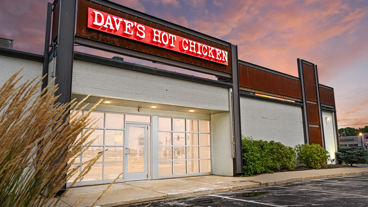 Exterior of Dave's Hot Chicken in Ashwaubenon, WI, near Green Bay, featuring a red neon sign and modern entrance at sunset.