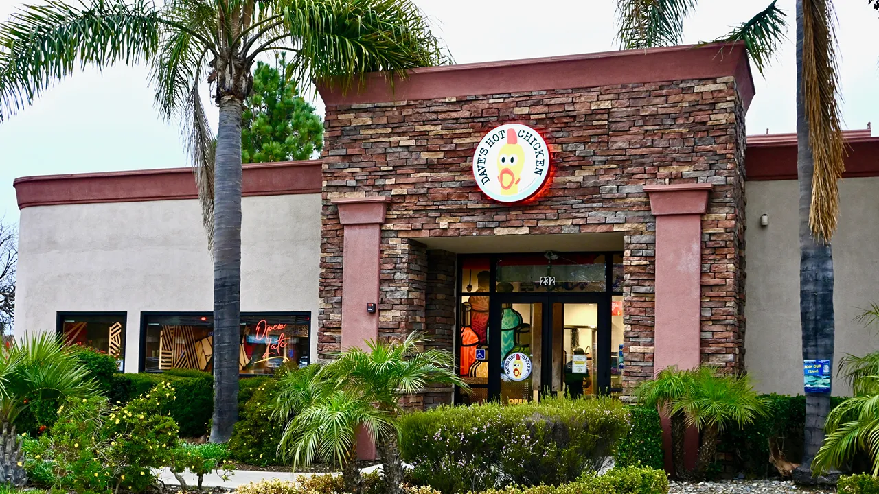 Exterior view of Dave’s Hot Chicken entrance on Betteravia Rd, Santa Maria, CA, featuring brick facade, palm trees, and colorful artwork.