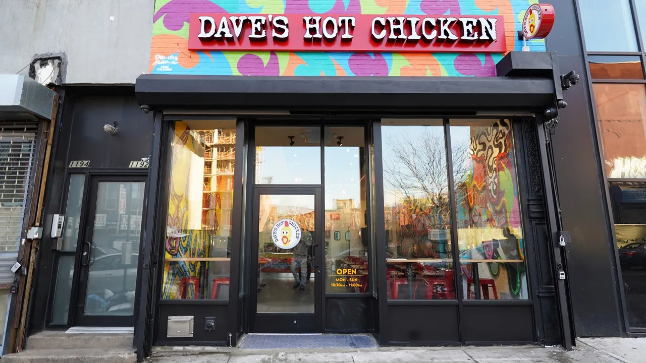 Exterior of Dave's Hot Chicken, Brooklyn, NY, on Fulton St with colorful graffiti art visible through the windows.