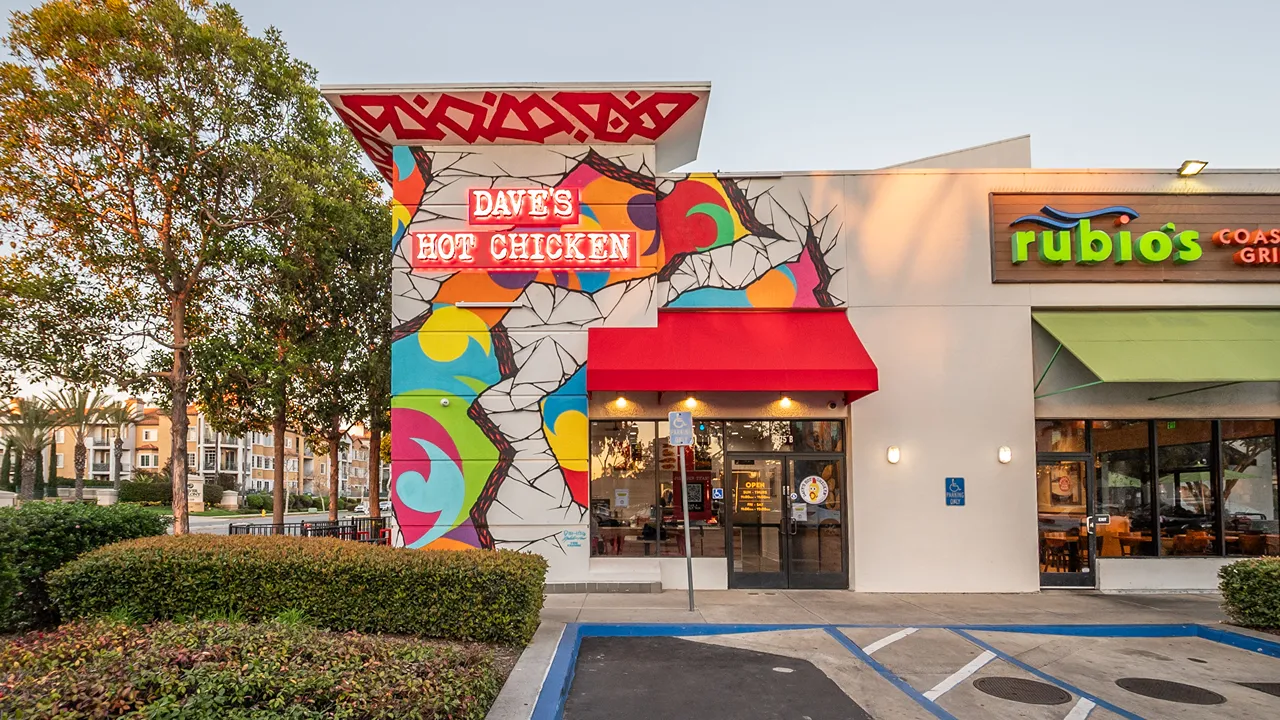 Side exterior view of Dave's Hot Chicken featuring a red awning and colorful mural at Camino De La Reina – Mission Valley, San Diego, CA.