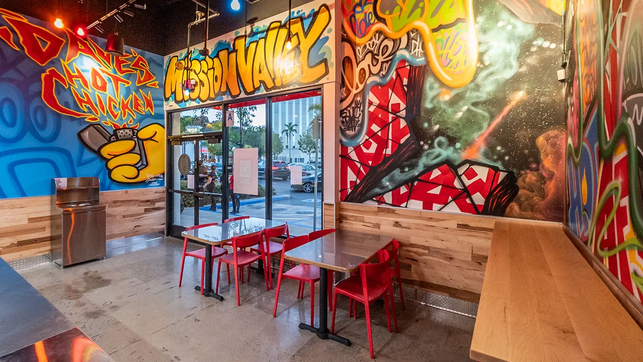 Seating area with red chairs and colorful mural featuring Mission Valley and bold graffiti designs inside Dave's Hot Chicken, Camino De La Reina – Mission Valley, San Diego, CA.