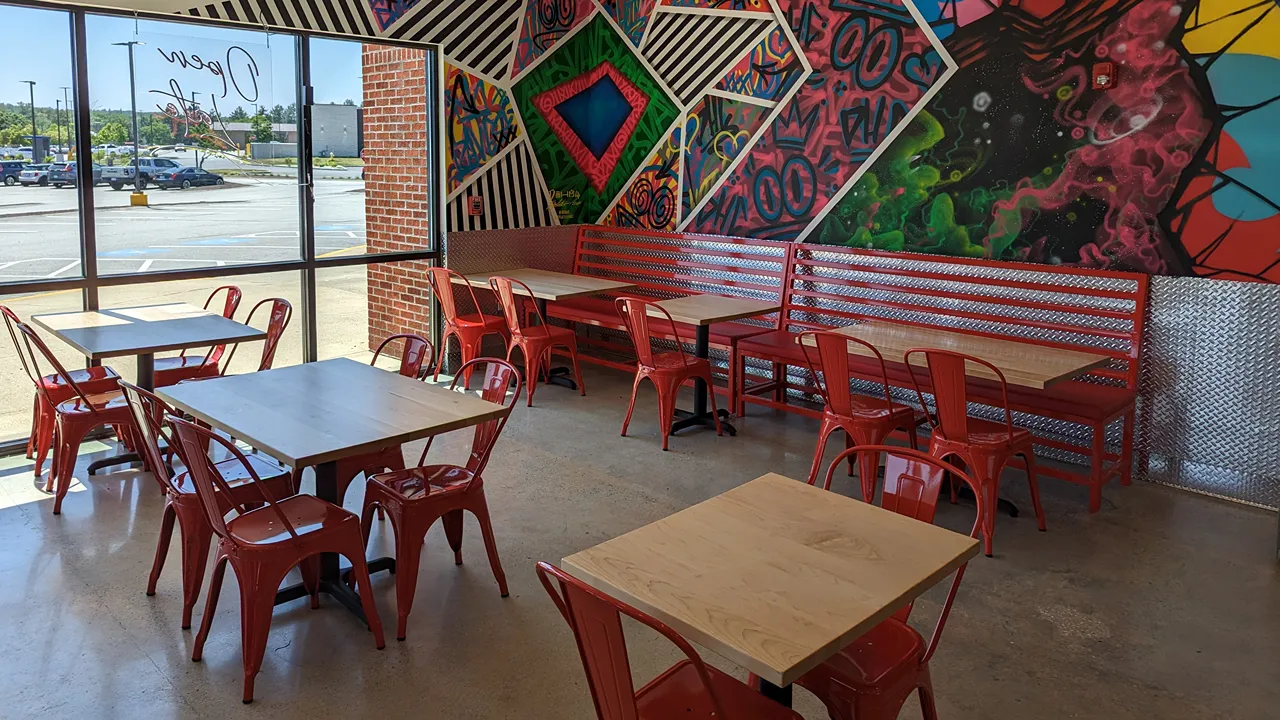 Interior seating of Dave's Hot Chicken, Chelmsford, MA with red chairs, wooden tables, and large window views with colourful wall murals.