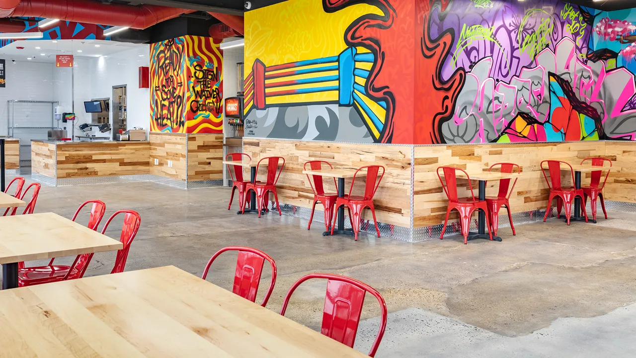 A Spacious Dave's Hot Chicken Restaurant in Cherry Hill, NJ, Ellisburg showing 3 graffiti murals covering 3 different walls interconnected by vibrant colours with tables and bright red chairs.