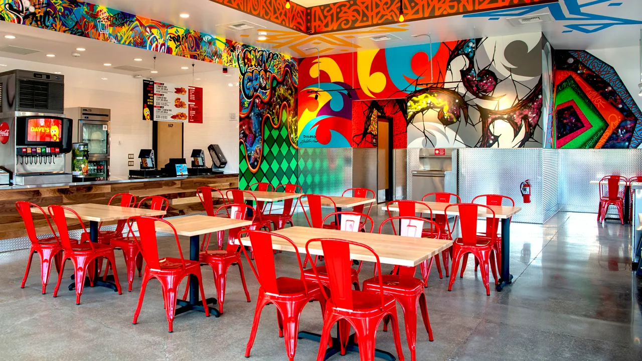 Red chairs and wooden tables with colorful murals on the walls inside Dave's Hot Chicken in Clackamas, OR.