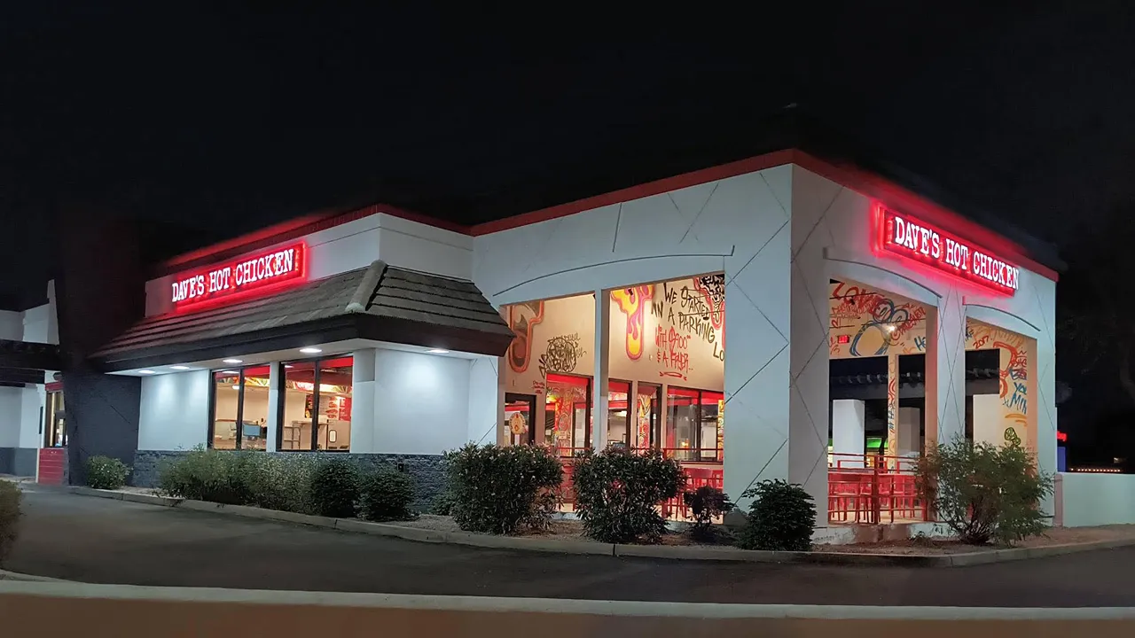 Night view of the exterior of Dave’s Hot Chicken in Gilbert, AZ with red signage glowing and graffiti art visible through large windows.