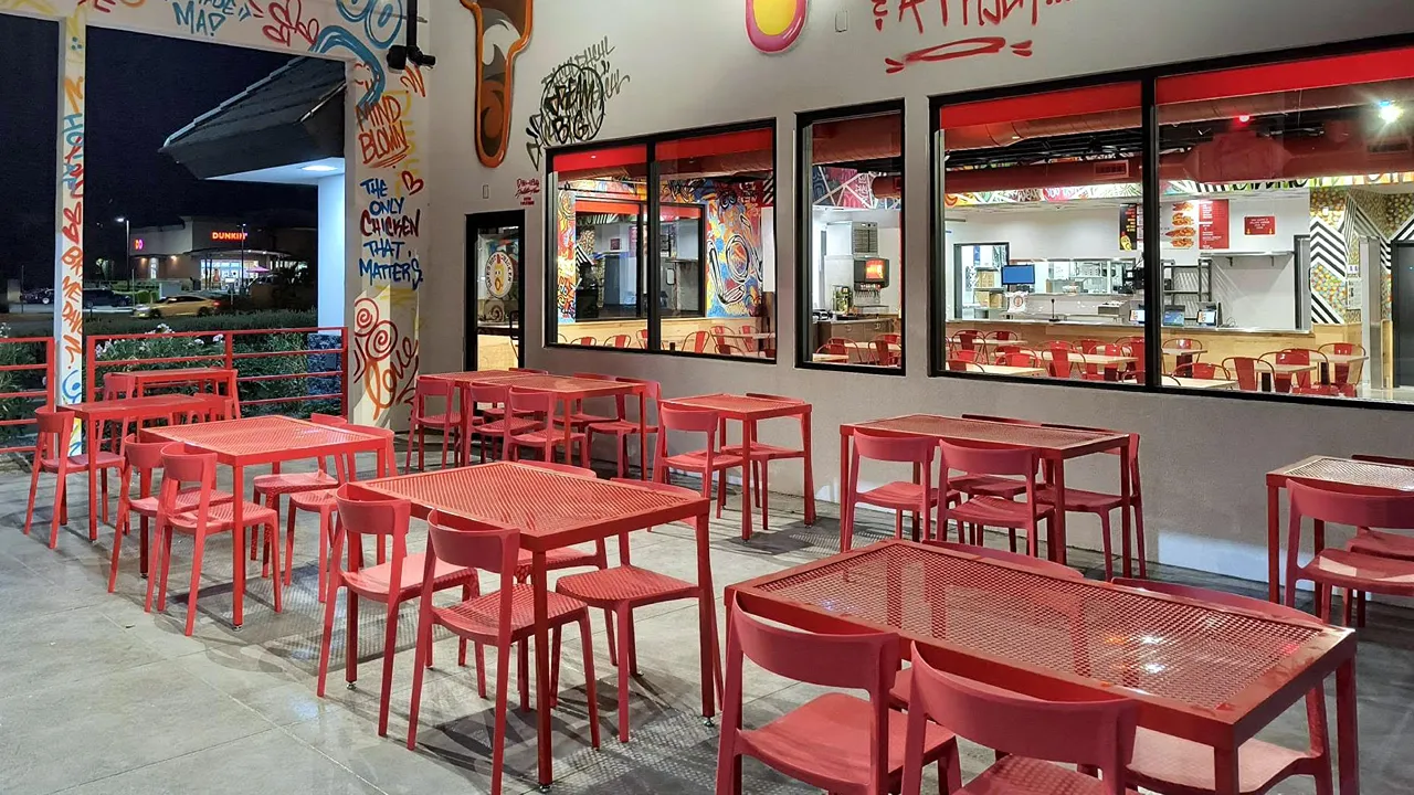 Outdoor seating area with red metal tables and chairs, featuring a view of the interior through large windows at Dave’s Hot Chicken in Gilbert, AZ.