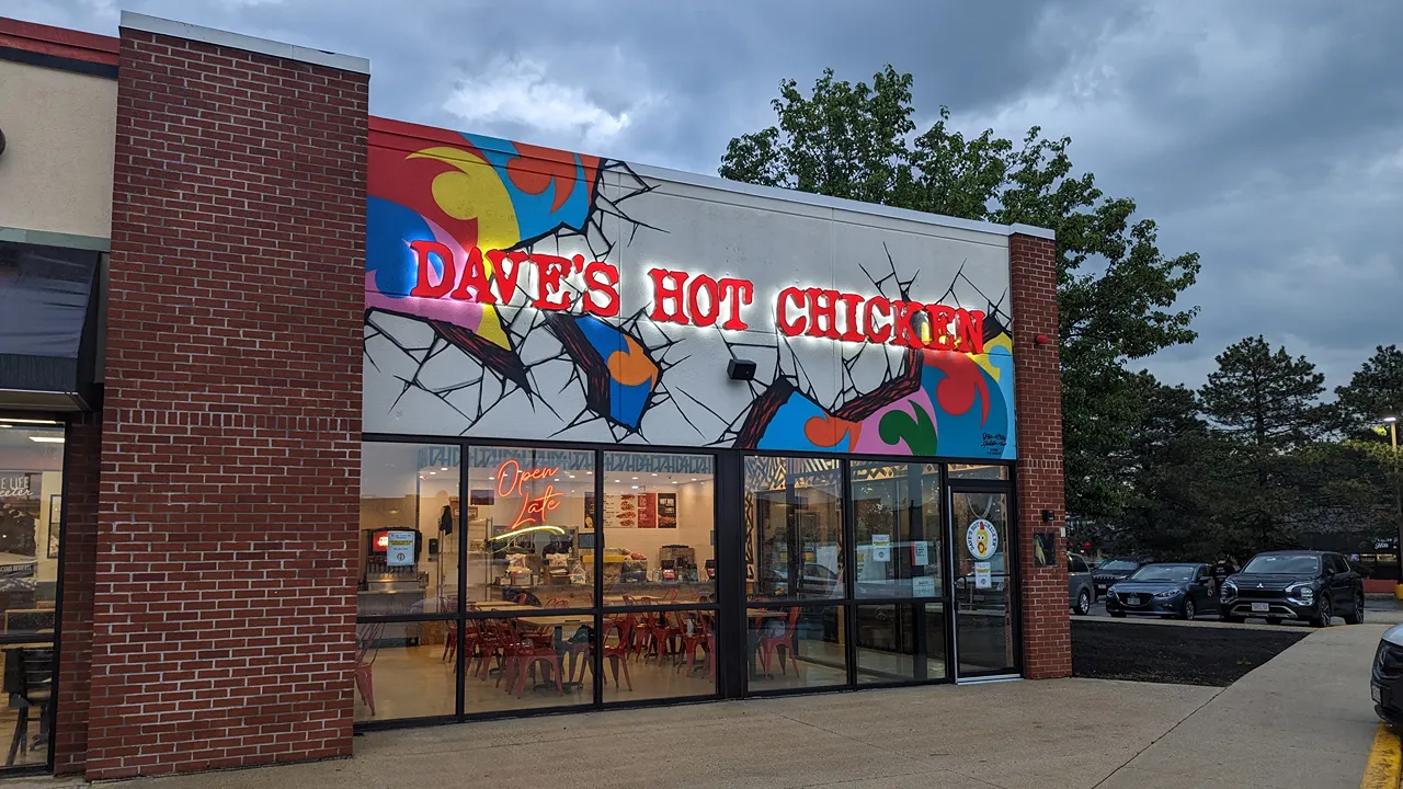 View from outside Dave's Hot Chicken Chelmsford, MA with Dave's colorful abstract signage. There is a view through windows showing the counter and seating area.