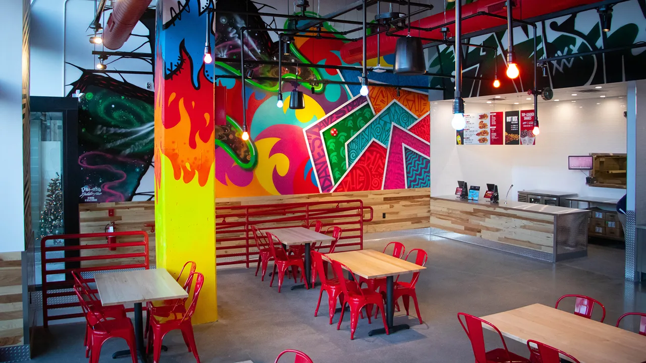 Bright and colorful wall murals with abstract designs and red chairs surrounding wooden tables in the seating area of Dave's Hot Chicken, East Lansing, MI.