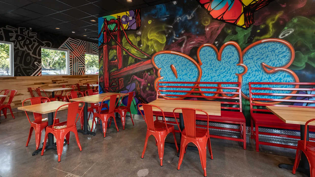 Mural featuring the Golden Gate Bridge and seating area with red chairs at Dave's Hot Chicken in El Cerrito Plaza, El Cerrito, CA.