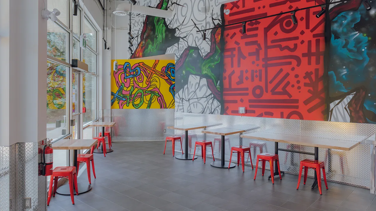 Interior of Dave's Hot Chicken in Burlington, ON, showing vibrant graffiti walls, tables with red stools, and street-facing windows.