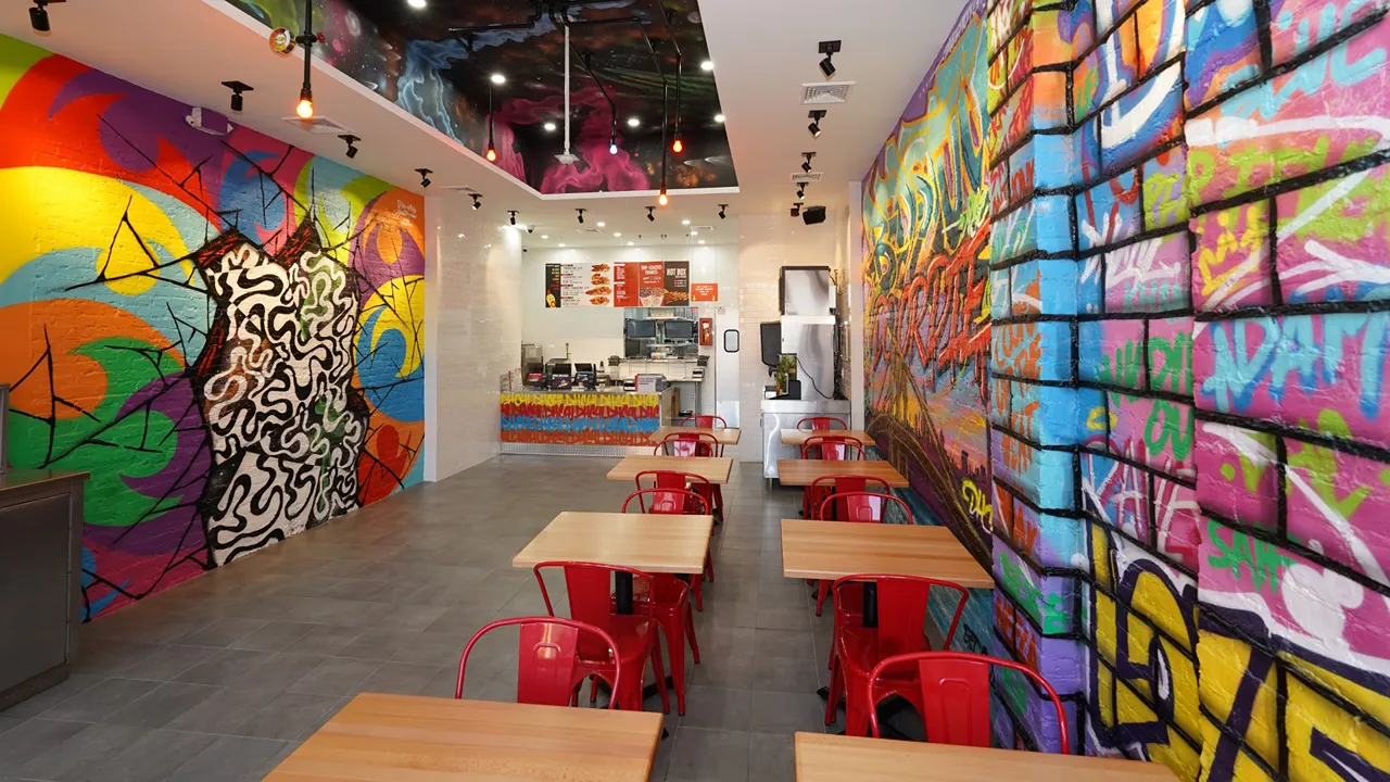 Interior of Dave's Hot Chicken on Fulton St, Brooklyn, NY, with graffiti art and red chairs surrounding wooden tables.