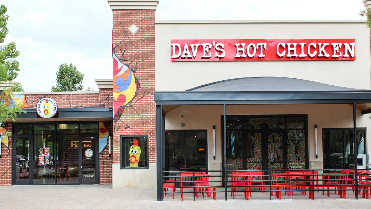 Daytime front view of Dave's Hot Chicken with red outdoor seating and large signage on Johnny Bench Dr., Oklahoma City, OK.