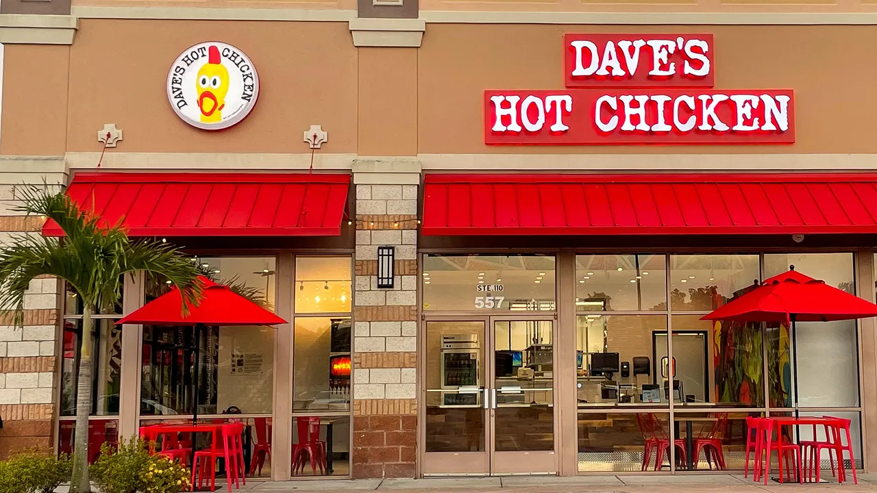 Front view of Dave's Hot Chicken on N. Alafaya Trail, Waterford Lakes, Orlando, FL, featuring outdoor seating with red umbrellas.