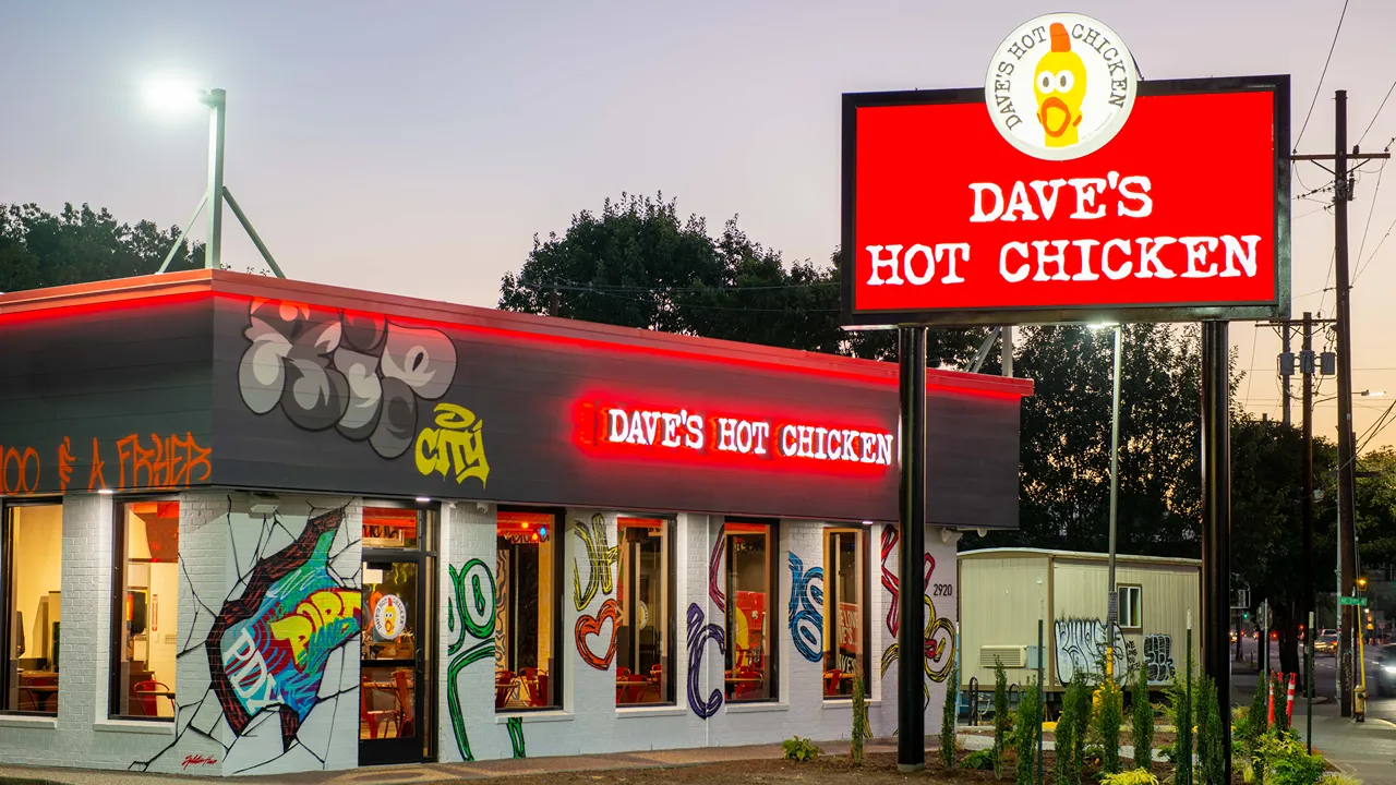 Exterior night view of Dave's Hot Chicken on NE Sandy Blvd, Portland, OR, with large red signage and graffiti artwork on the walls.