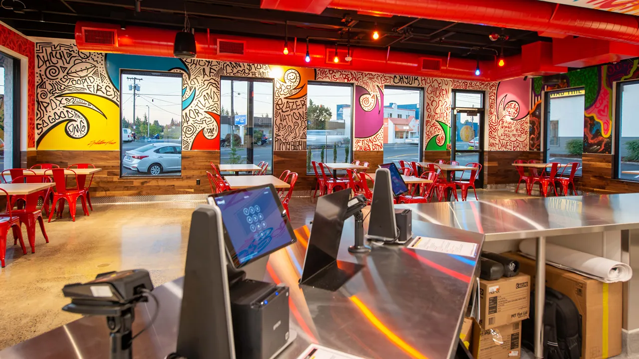 Indoor seating area at Dave's Hot Chicken on NE Sandy Blvd, Portland, OR, with colorful murals and red chairs around wooden tables.