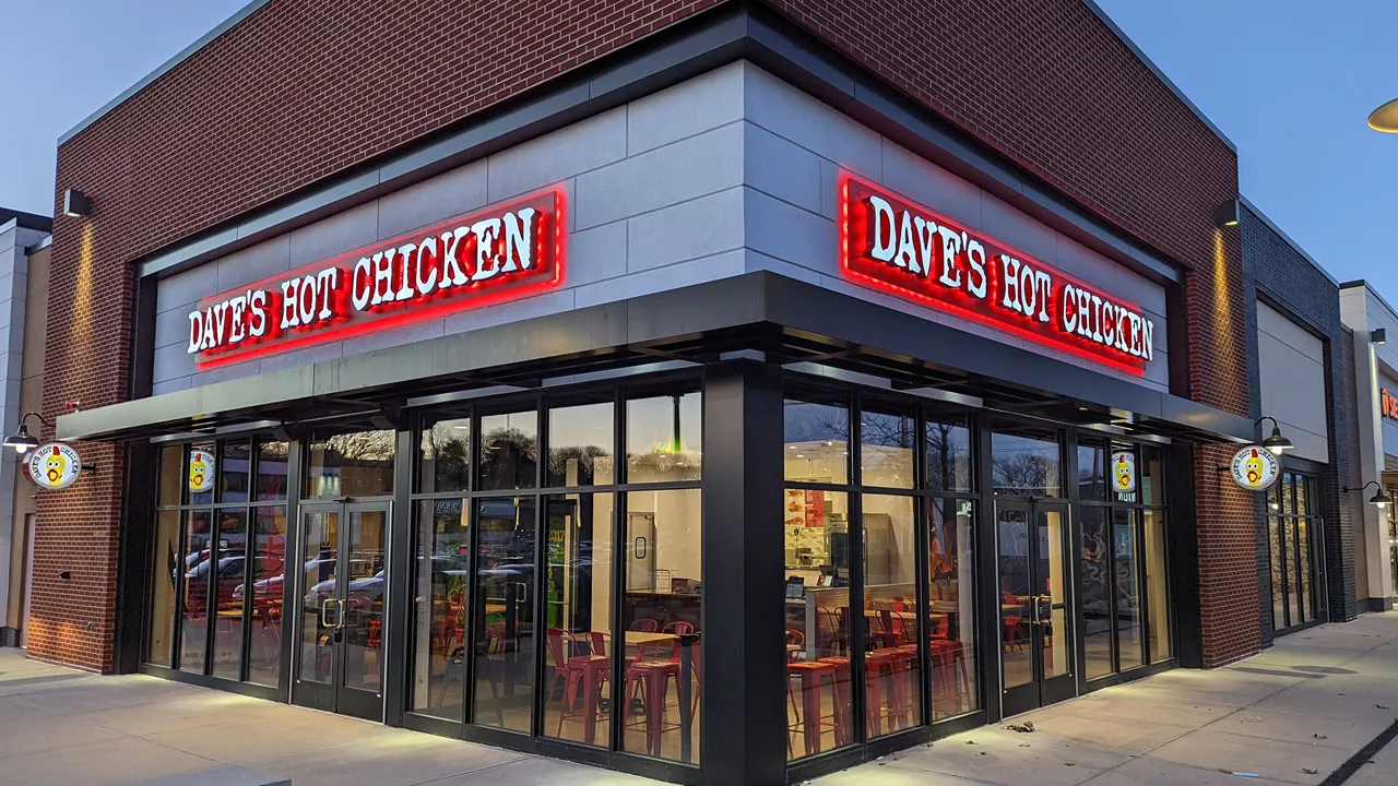 Night view of outside Dave’s Hot Chicken with bright signage and windows all around the building on Needham St., Newton, MA.