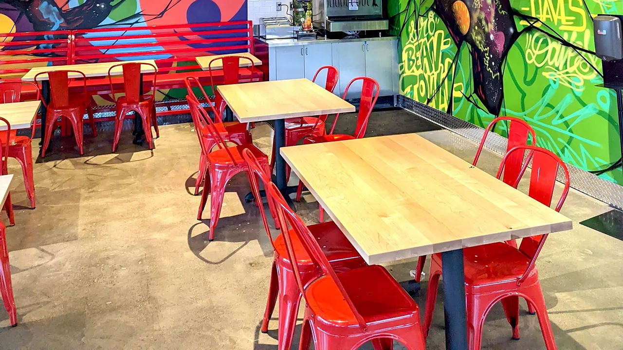 Close-up of red chairs and tables near vibrant murals in the seating area at Dave's Hot Chicken on Needham St., Newton, MA.