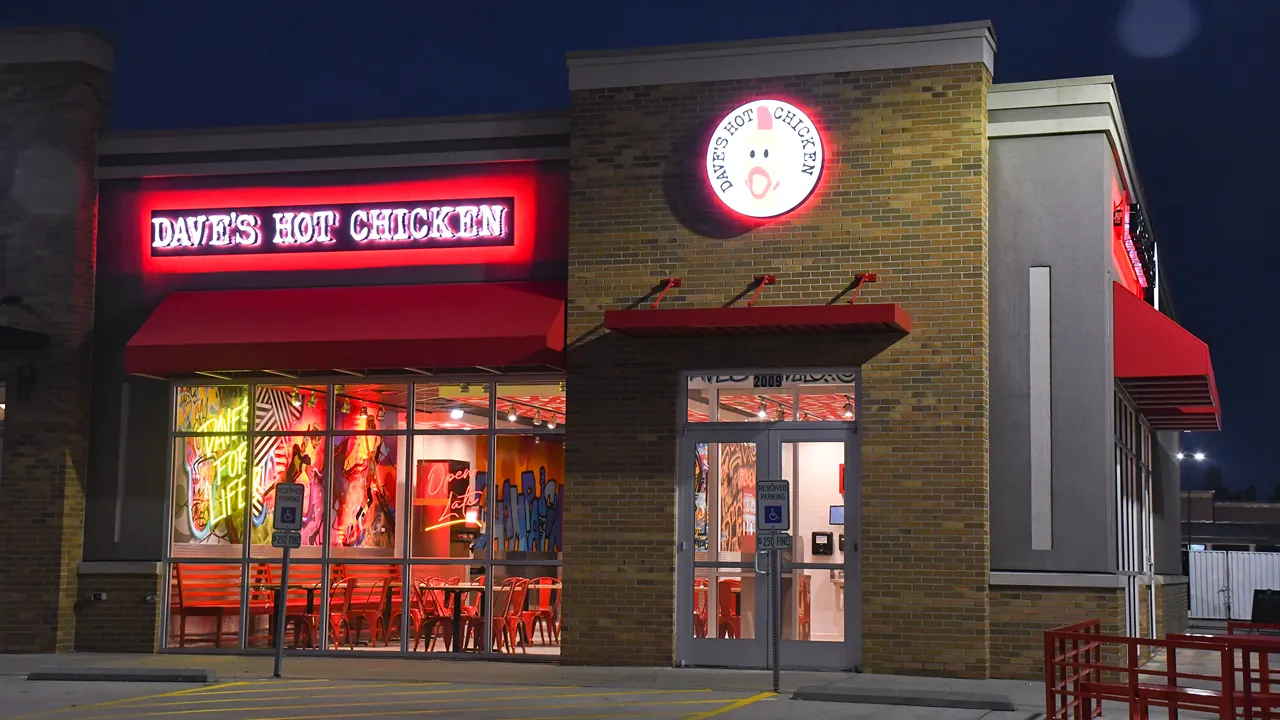 Exterior of Dave's Hot Chicken on Neil St, Champaign, IL, showing the building at night with red signage and seating inside.