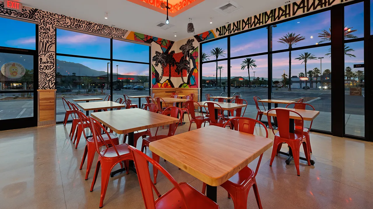 Interior of Dave's Hot Chicken in Cathedral City, CA, with a palm mural, red chairs, and wooden tables at sunset.