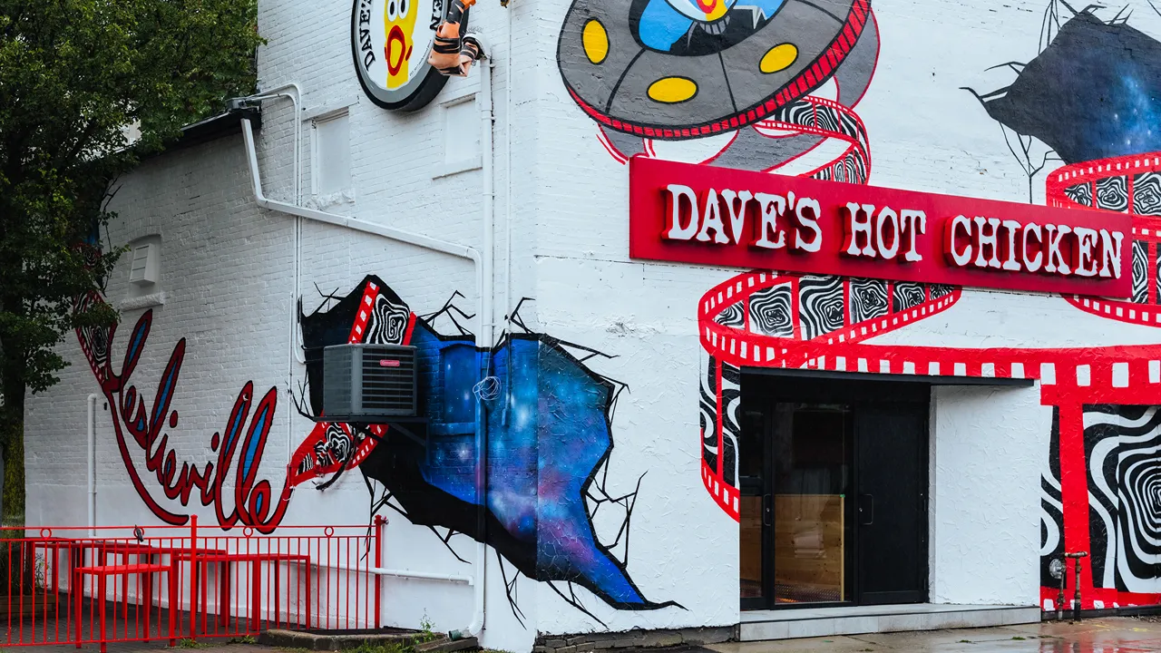 Dave's Hot Chicken storefront on Queen St E in Leslieville, Toronto, ON, with a colorful mural on the white building with the words 'Leslieville'.