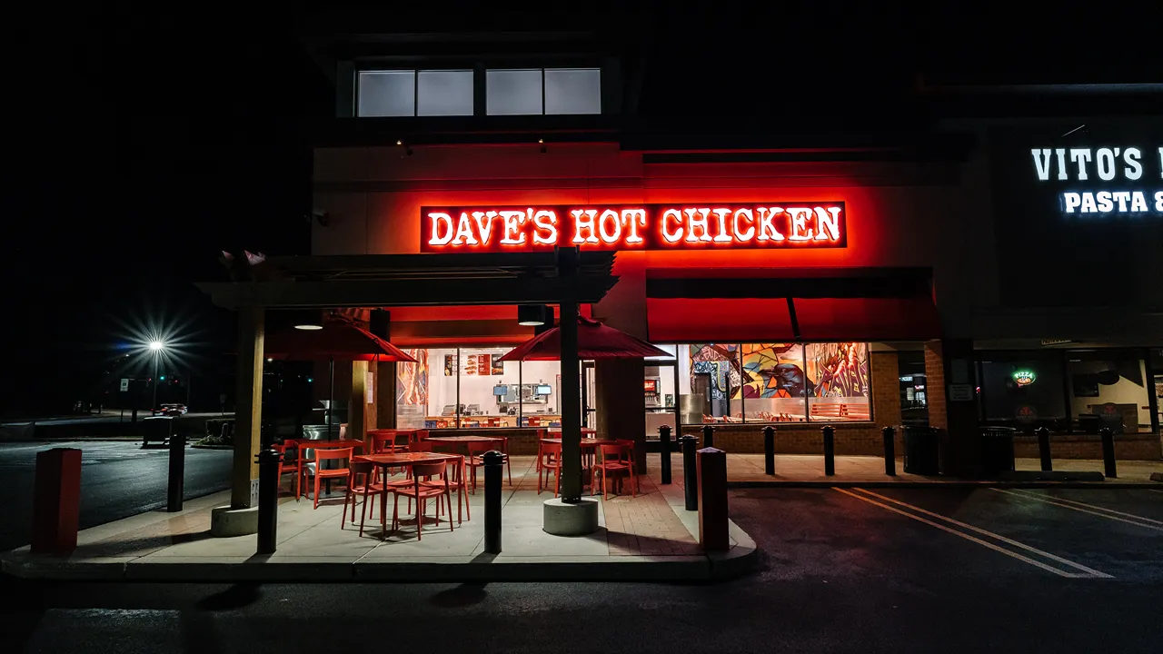 Nighttime view of the illuminated exterior of Dave’s Hot Chicken, showcasing outdoor seating and vibrant signage on Reisterstown Rd, Owings Mills, MD.