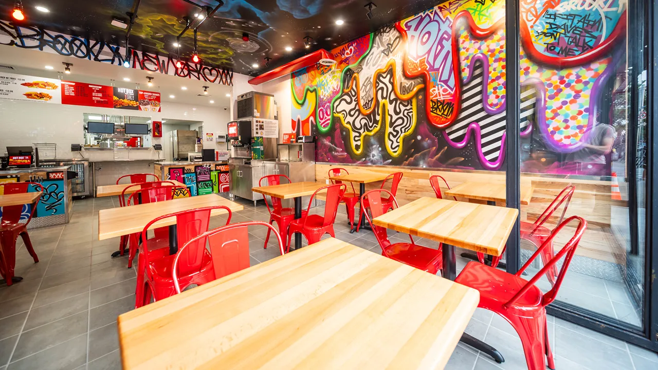 A wide view of Dave's Hot Chicken in Brooklyn, NY, highlighting the graffiti-covered walls, red chairs, wooden tables, and the service counter.