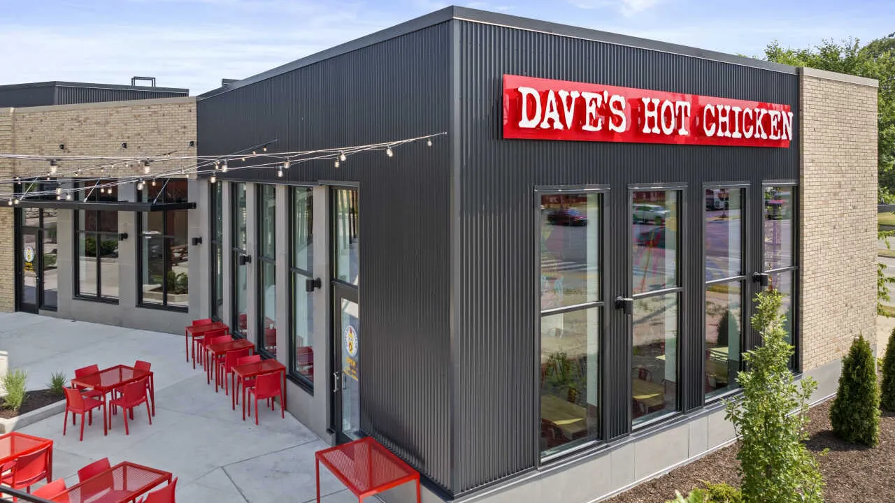 Corner View of the Dave's Hot Chicken Restaurant on Sligo Drive, Madison, WI, featuring signage, large windows with red chairs and tables under string lights.