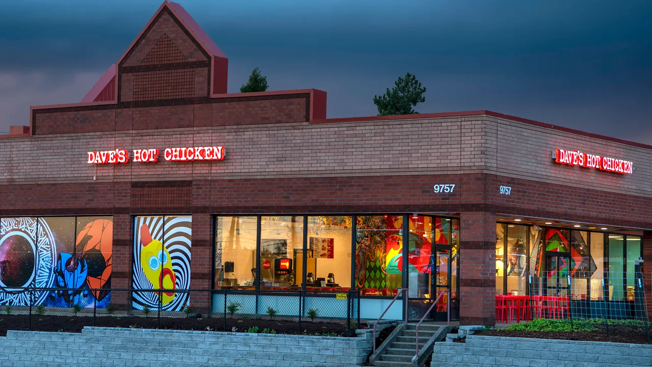 Óutside Dave's Hot Chicken restaurant in Clackamas, OR with large windows, showing bright murals from inside, and illuminated signage.