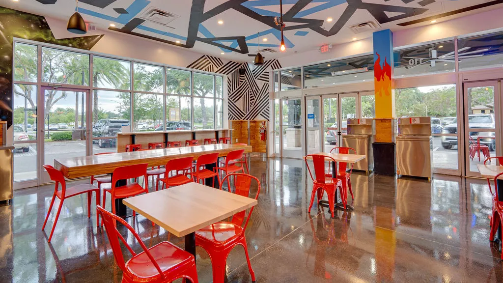 Bright seating area with red chairs and tables, large windows, and colorful wall art inside Dave’s Hot Chicken restaurant in Gainesville, FL