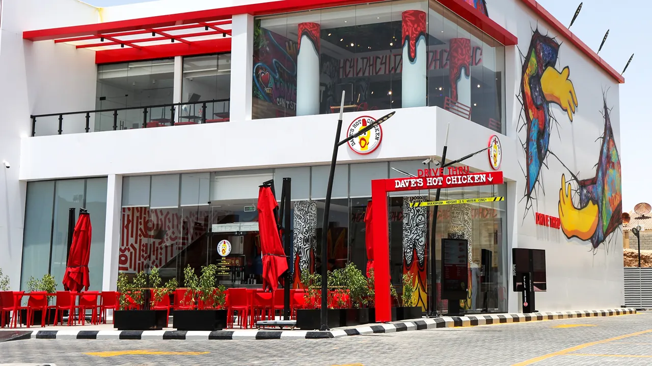Drive-thru entrance at Dave's Hot Chicken on Uthman Ibn Affan Branch Rd, Riyadh, KSA, with outdoor seating and graffiti art.