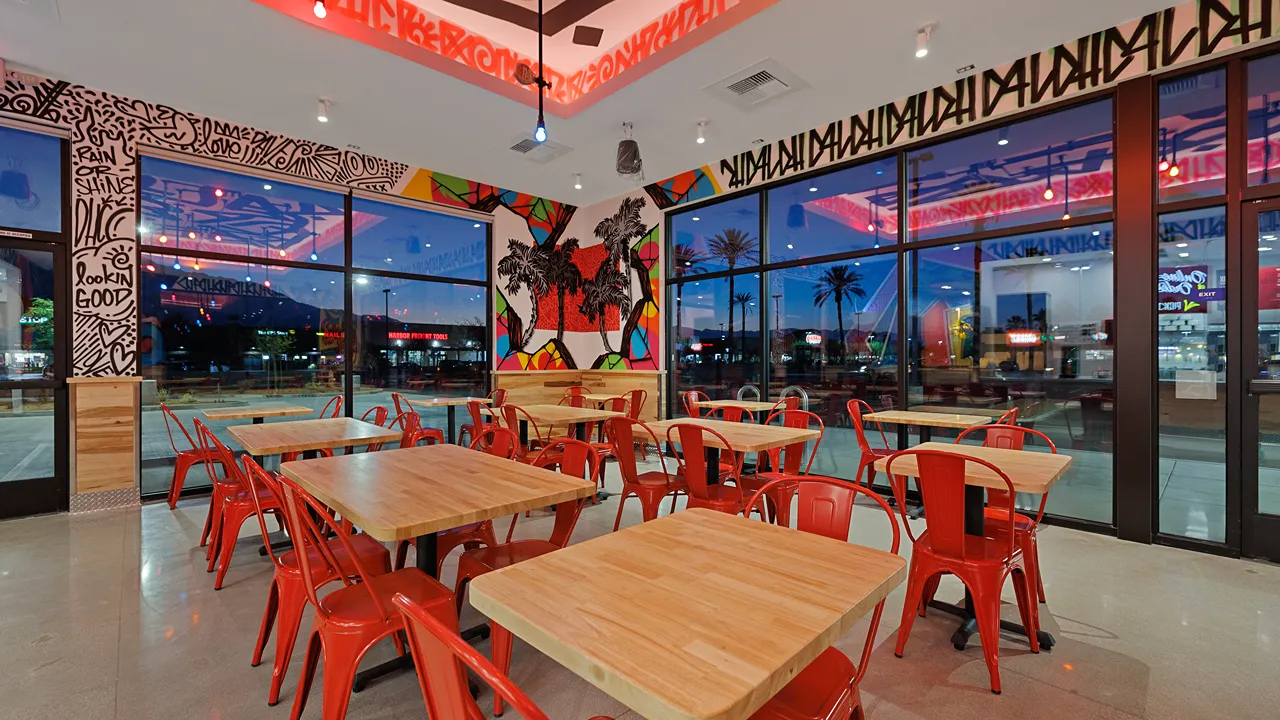 Evening view from inside Dave's Hot Chicken in Cathedral City, CA, showing red chairs and tables with large windows and a sunset.